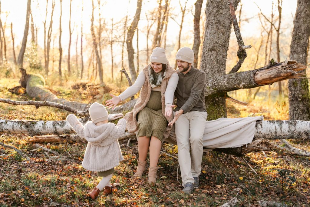 Schwangere Mama und Papa sitzen auf einem Baumstamm, ihre Tochter rennt mit ausgestreckten Armen auf sie zu