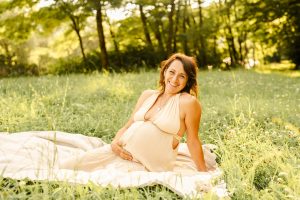 Schwangere im beigen Kleid sitzt entspannt auf einer Decke auf einer Wiese und lächelt in die Kamera