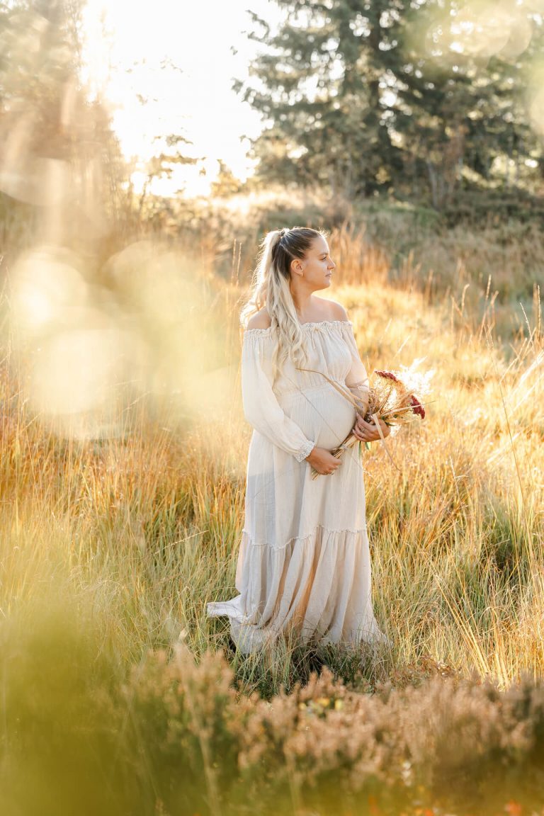 Schwangere steht auf einer Wiese im Sonnenunterganslicht, in den Armen vor dem Babybauch hält sie einen Blumenstrauß