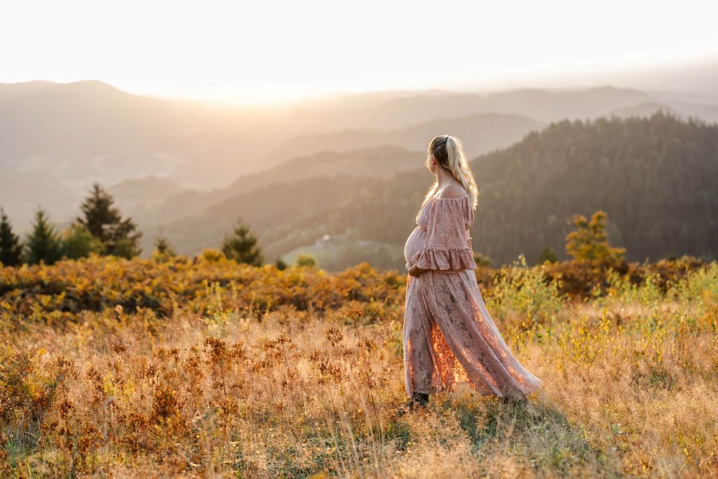 Schwangere in altrosafarbenen Spitzenkleid läuft auf einer Wiese und betrachtet die Schwarzwaldberge im Sonnenuntergangslicht