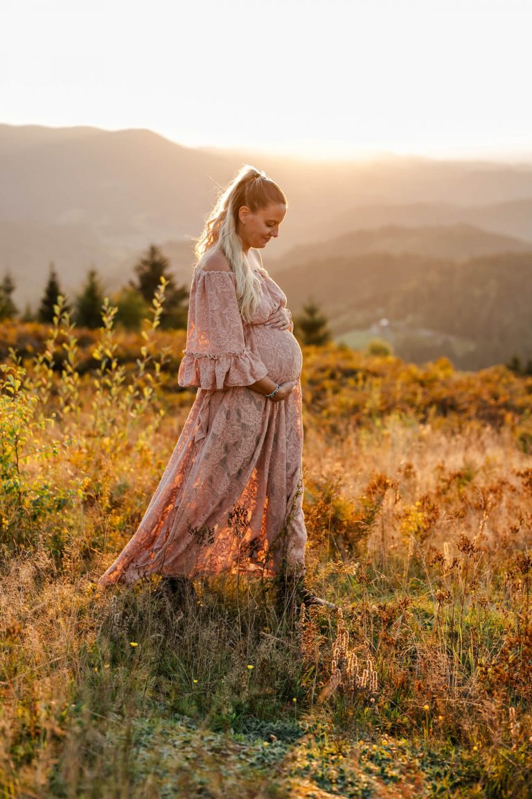 Schwangere in altrosa Spitzenkleid läuft über eine Wiese im Sonnenuntergang, im Hintergrund sind Schwarzwaldberge
