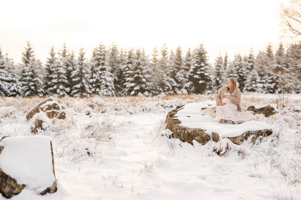 Schwangere sitzt auf einem großen Stein in schöner Schneelandschaft, verschneite Bäume im Hintergrund