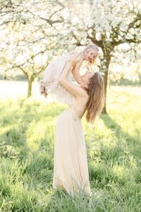 Schwangere im beigen Kleid steht auf einer Wiese und hebt ihre Tochter in die Luft, beide lachen