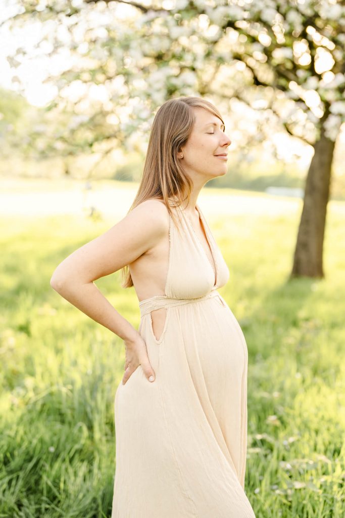 Schwangere steht mit geschlossenen Augen auf Wiese im Sonnenuntergangslicht