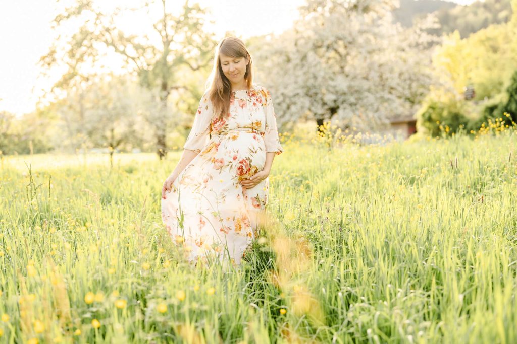 Schwangere im geblümten Kleid läuft über eine Wiese und schwingt ihr Kleid