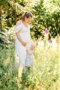 Schwangere im weißen Spitzenkleid steht auf einer Wiese mit Blumen, ihre kleine Tochter kuschelt sich an sie und beide lachen