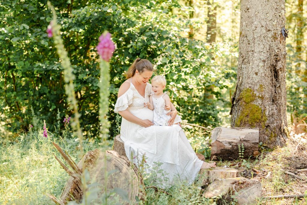 Schwangere Mama sitzt im Wald auf einem Baumstumpf, ihre kleine Tochter ist neben ihr, beide lachen