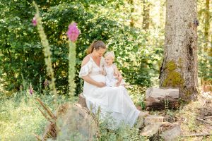 Schwangere im Spitzenkleid sitzt auf einem Baumstumpf im Wald mit ihrer kleinen Tochter auf dem Schoß, beide lachen
