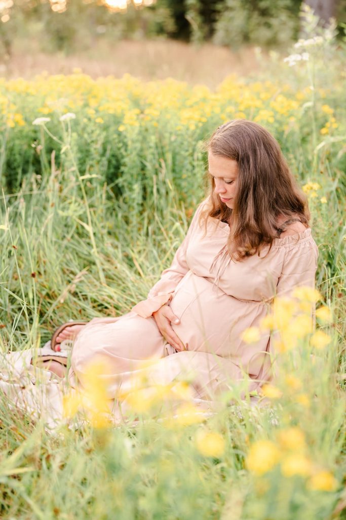 Schwangere sitzt in einer Wiese mit gelben Blumen und hält die Hand an ihren Babybauch