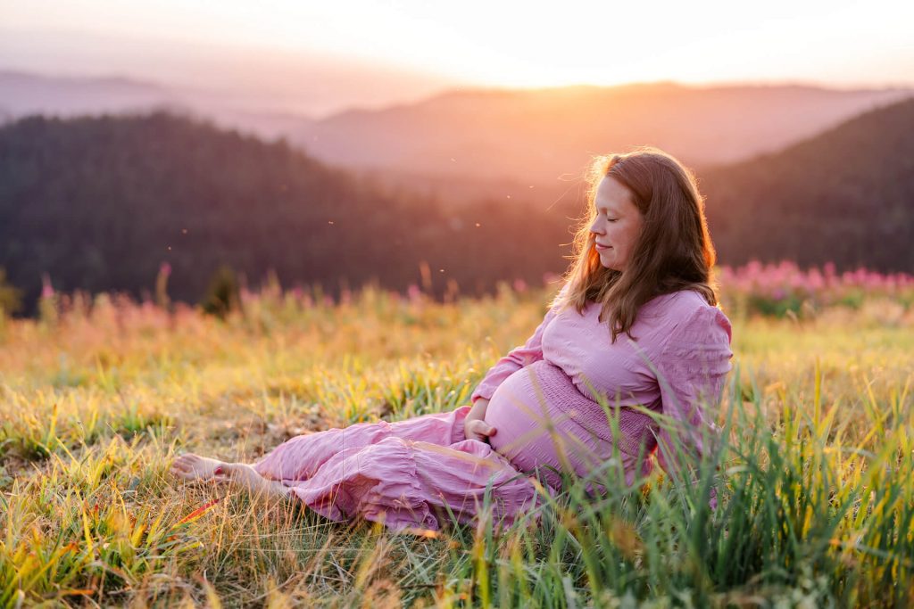 Schwangere sitzt mit geschlossenen Augen auf einer Wiese, im Hintergrund Schwarzwaldberge im Sonnenuntergang