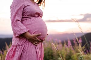 Schwangere im hübschen Altrosa Kleid steht auf einer Wiese und streichelt über ihren Babybauch