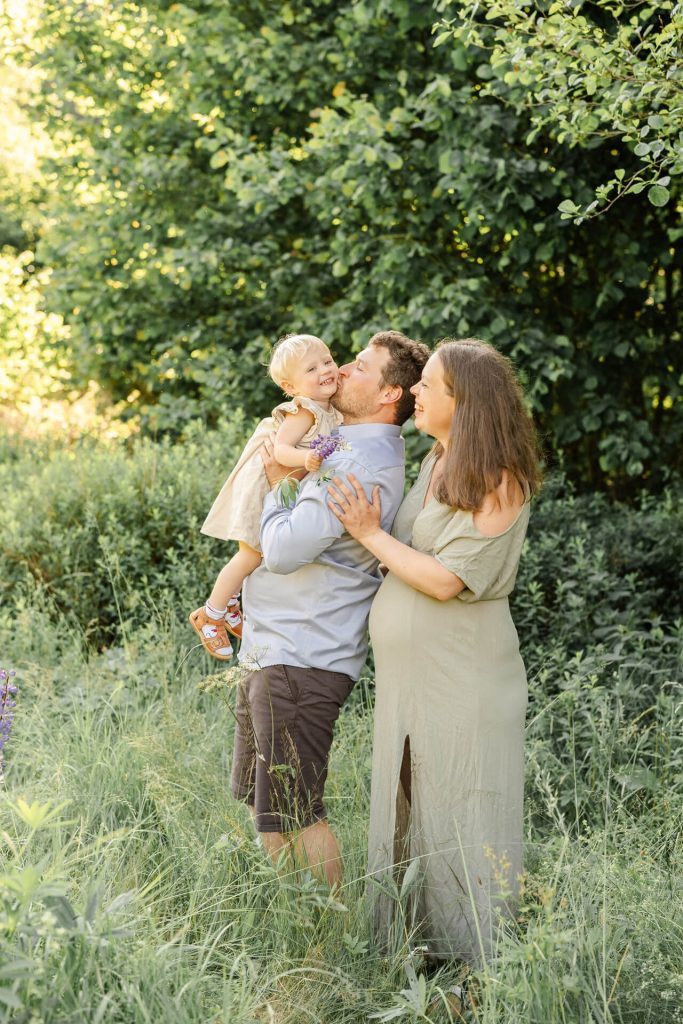 Schwangere Mama und Papa stehen auf einer Wiese, Papa hält die Tochter in den Armen und gibt ihr einen Kuss auf die Wange