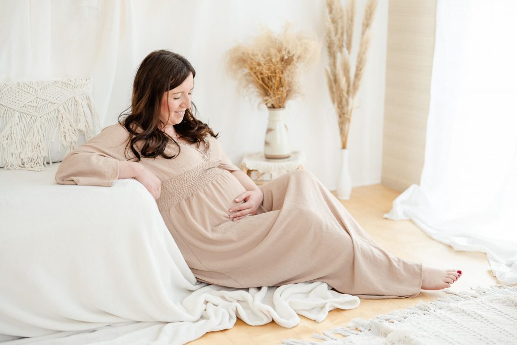 Schwangere sitzt gemütlich auf dem Boden und lehnt sich an ein Sofa, betrachtet ihren Babybauch