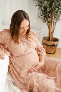 Schwangere im altrosa Spitzenkleid lehnt sich gemütlich an ein Sofa und streicht über ihren Bauch