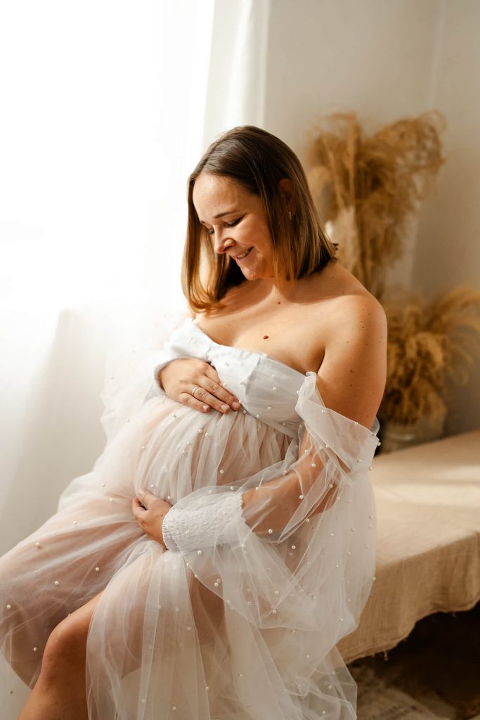 Schwangere in Tüllkleid sitzt auf einer Bank, streicht und lächelt ihren Babybauch an