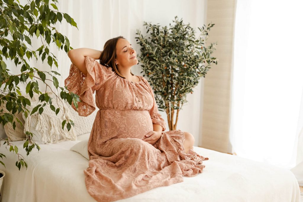 Schwangere in schönem Spitzenkleid sitzt auf Sofa, streicht über ihren Bauch und hat eine Hand in den Haaren