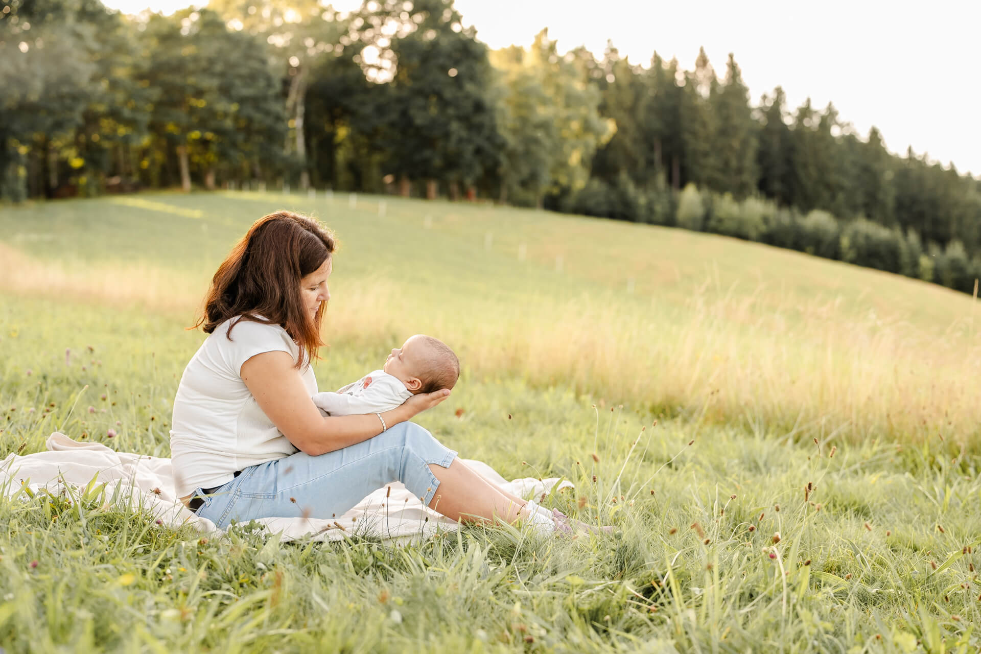 Mama sitzt auf einer Decke auf der Wiese und hält ihr Neugeborenes auf den Oberschenkeln in den Armen und betrachtet es