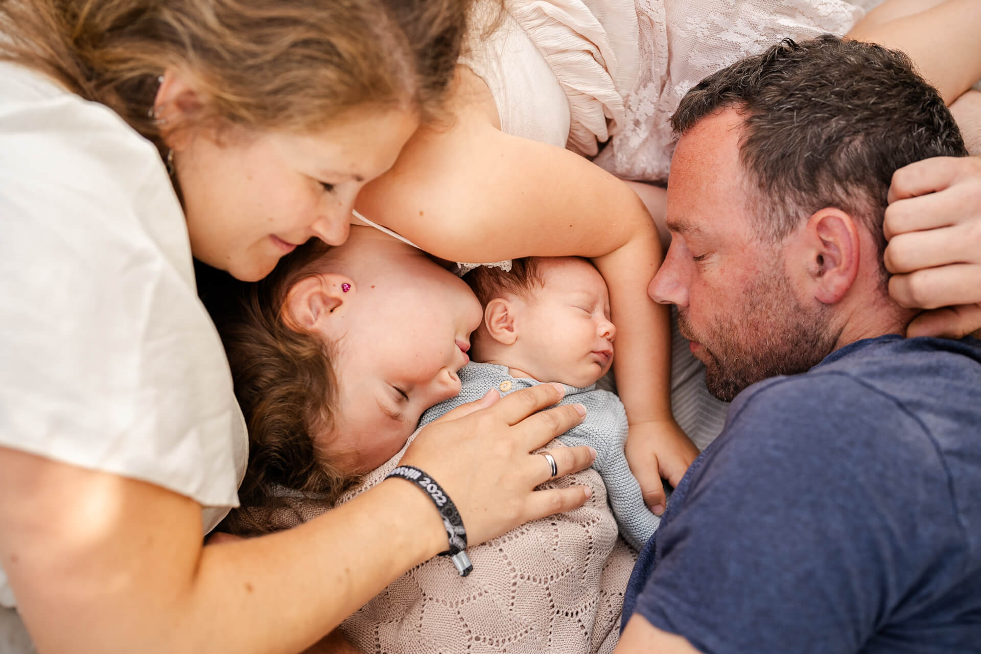 Mama und Papa liegen mit ihrem Neugeborenen und der großen SChwester eng aneinandergekuschelt, alle haben die Augen geschlossen