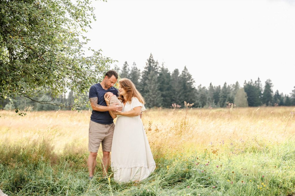 Mama und Papa stehen auf einer Wiese, mit Bäumen im Hintergrund, Sie halten ihr Neugeborenes in den Armen und betrachten es voller Liebe