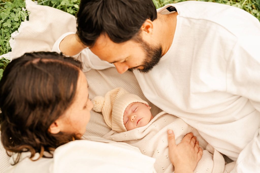 Mama und Papa liegen auf einer Decke auf einer Wiese, ihr Neugeborenes liegt eingekuschelt zwischen ihnen , sie betrachten es voller Liebe