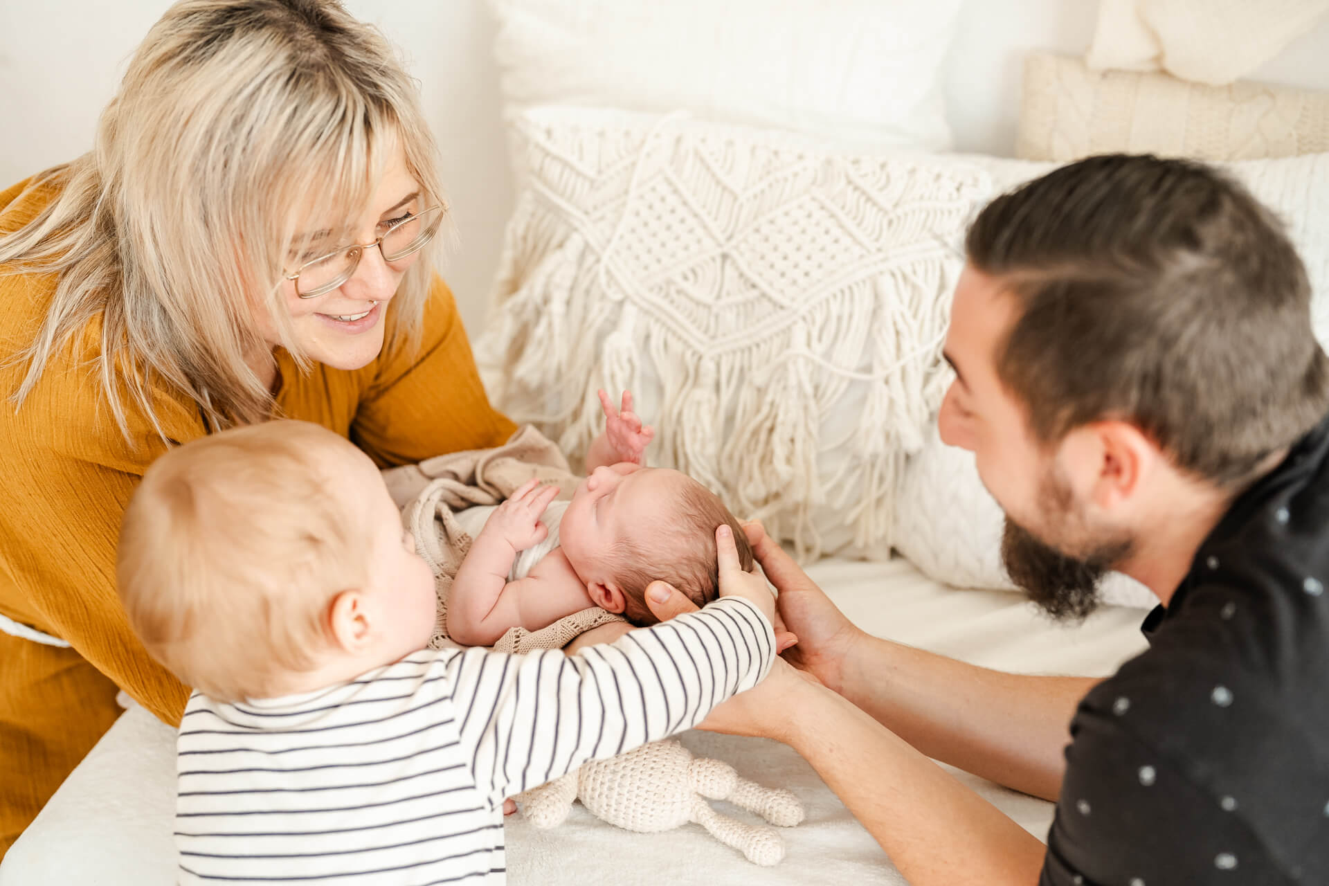 Mama und Papa sind sich gegenüber und halten ihr Neugeborenes in den Händen zwischen sich. Der große Bruder sitzt daneben und streichelt das Baby am Kopf, alle schauen zum Baby