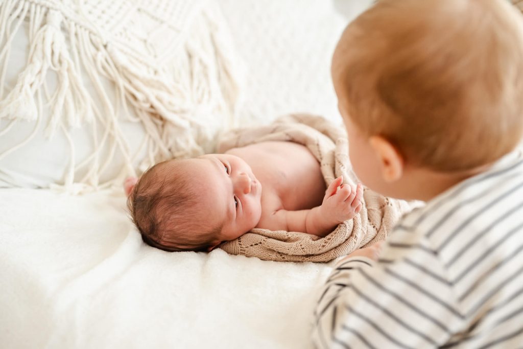 Neugeborenes liegt eingekuschelt in Decken auf einem Sofa, der große Bruder ist daneben unbd betrachtet es