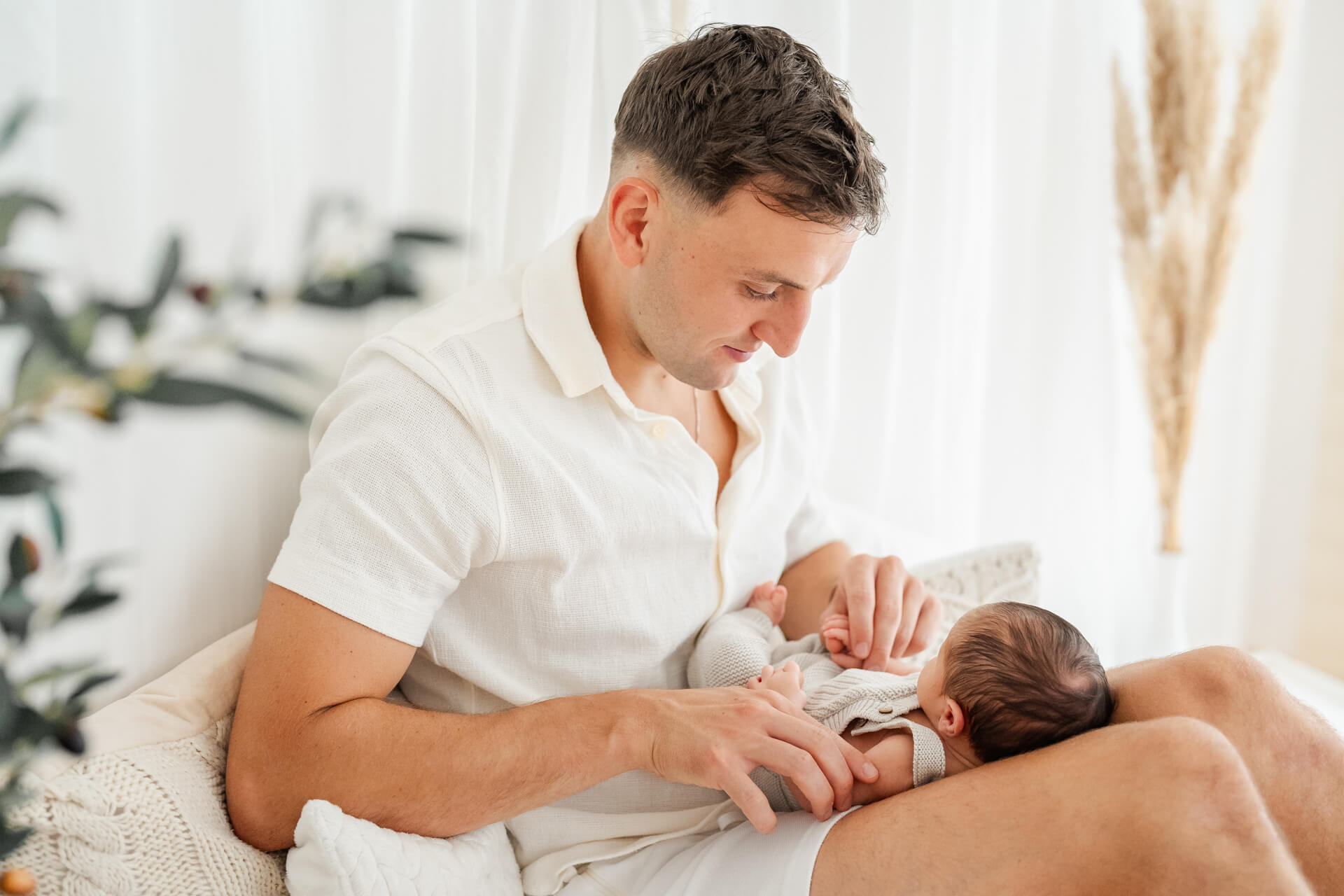 Papa sitzt mit Neugeborenem auf dem Sofa, es leit auf seinen Oberschenkeln, er betrachtet es