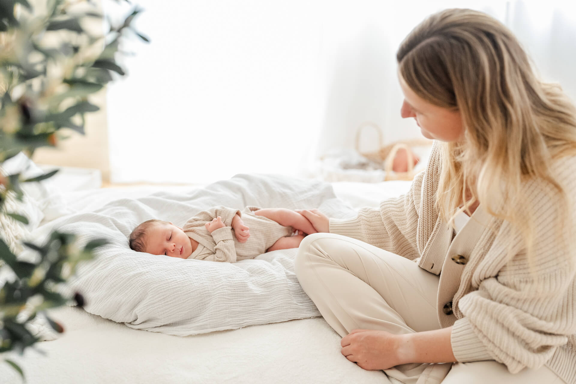 Neugeborenes liegt eingekuschelt in weiche Decken auf einem Sofa, die Mama sitzt daneben und betrachtet ihr Baby voll Liebe