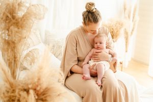 Mama im schönen beigen Kleid sitzt auf gemütlichem Sofa und hält ihr Baby im Arm