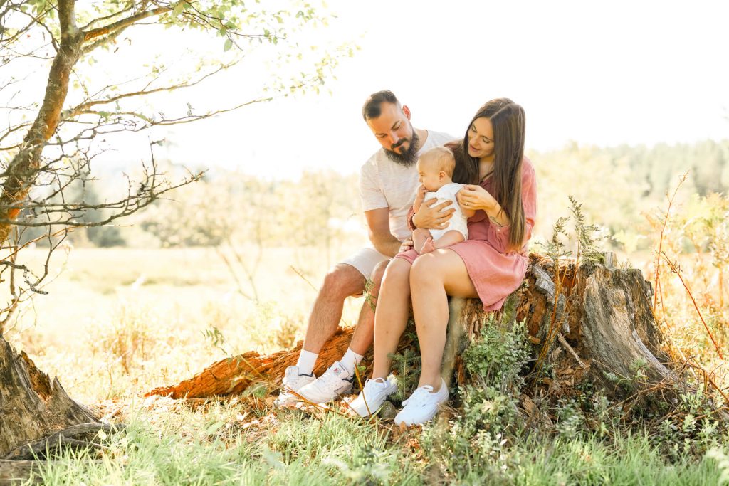 Familie, Mama, Papa und kleine Tochter sitzen zusammen auf einem Baumstumpf, kuscheln aneinander