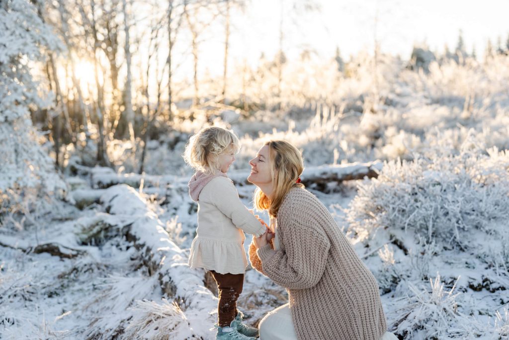 Mama und Tochter in einer Schneelandschaft, Das Mädchen steht auf auf einem Baumstamm, Mama kn iet vor ihr, sie halten sich an den Händen und lächeln sich an
