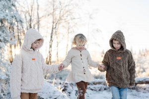 3 Kinder im Schnee, die beiden Jungs halten ihre kleine Schwester an den Händen, die über einen Baumstamm läuft