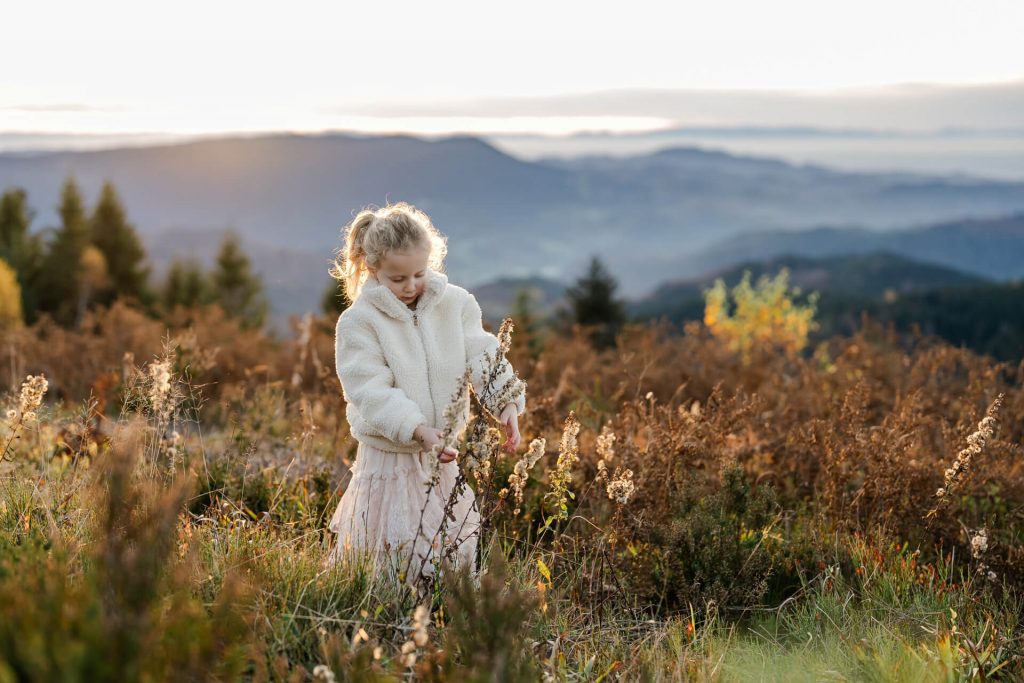 Mädchen steht auf einer WIese mit hohem Gras und schaut sich die Blumen an, im Hintergrund sind Schwarzwaldberge