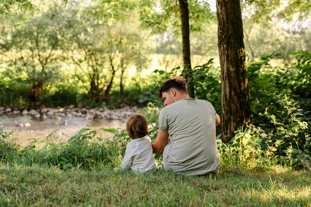 Papa und Sohn sitzen auf einer Wiese, Papa schaut zum Kind, der Junge schaut auf den Fluss