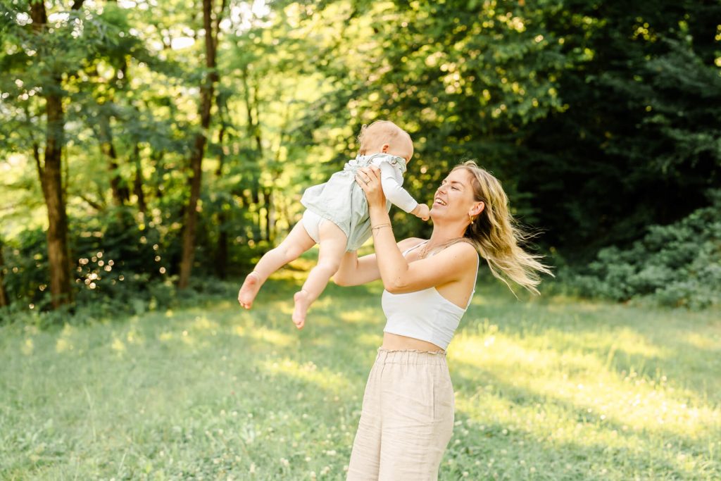 Mama schwingt ihre kleine Tochter in der Luft, sie lacht ihr Kind an. Sie steht auf einer Wiese, in Hintergrund sind Bäume im Sonnenuntergangslicht
