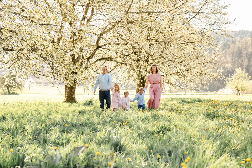 Familie (Mama, Papa und drei Kinder) halten sich an den Händen und laufen über eine Blumenwiese, Hinten sind blühende Kirschbäume