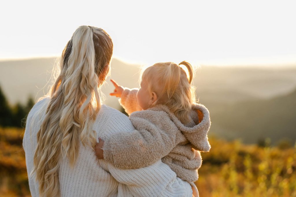 Mama hält ihre kleine Tochter auf dem Arm, diese streckt den Finger aus und zeigt Richtung Sonnenuntergang