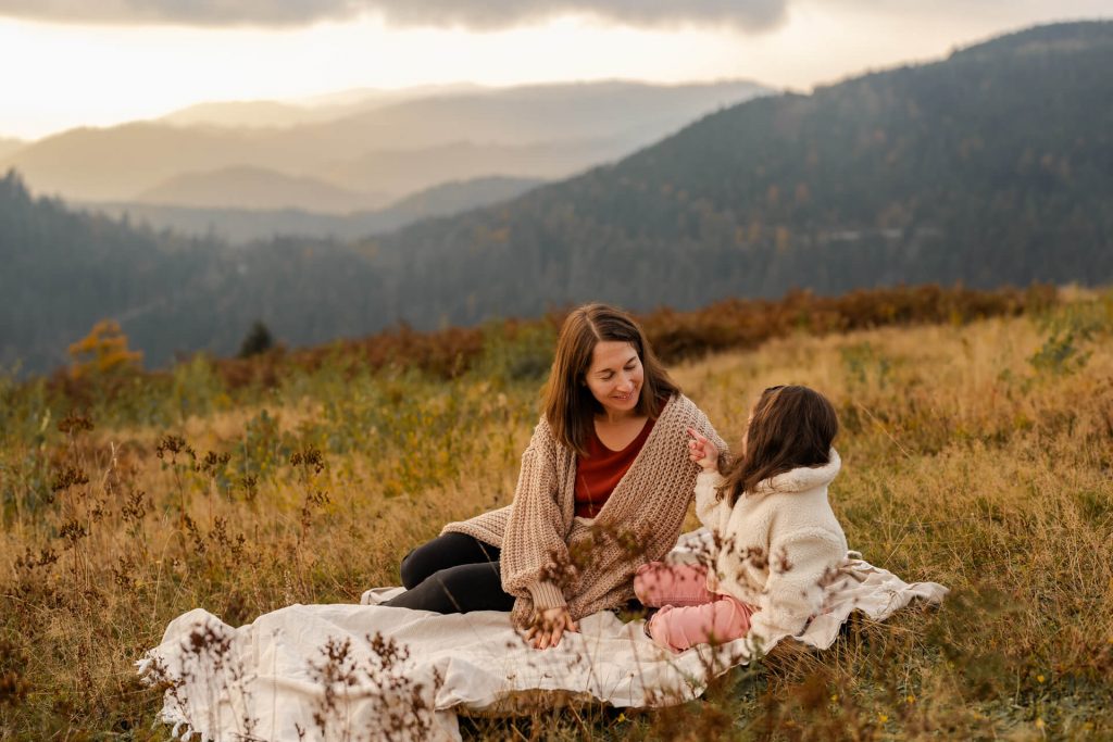 Mama und Tochter sitzen auf einer Decke und schauen sich an. IN HIntergrund sind Schwarzwaldberge im Sonnenuntergang