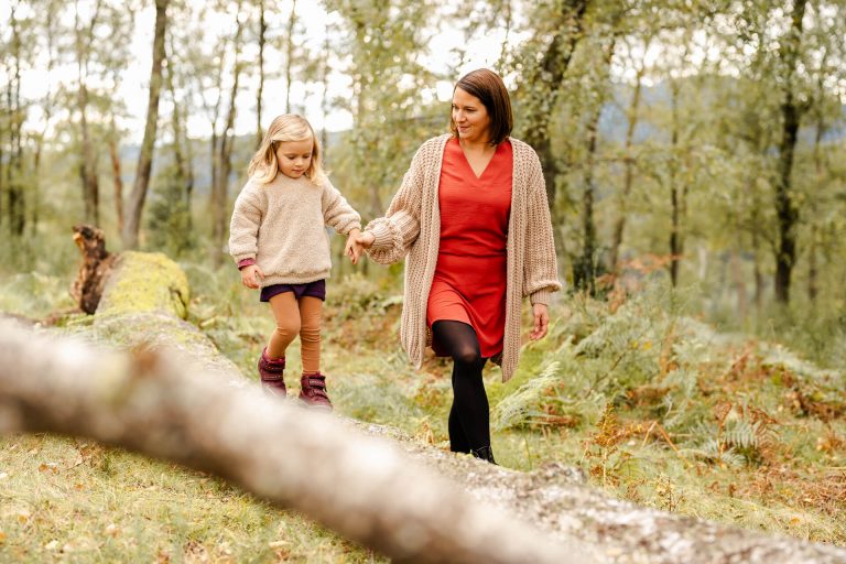 Mama hält ihre Tochter an der Hand, die über einen Baumstamm balanciert, beide lachen