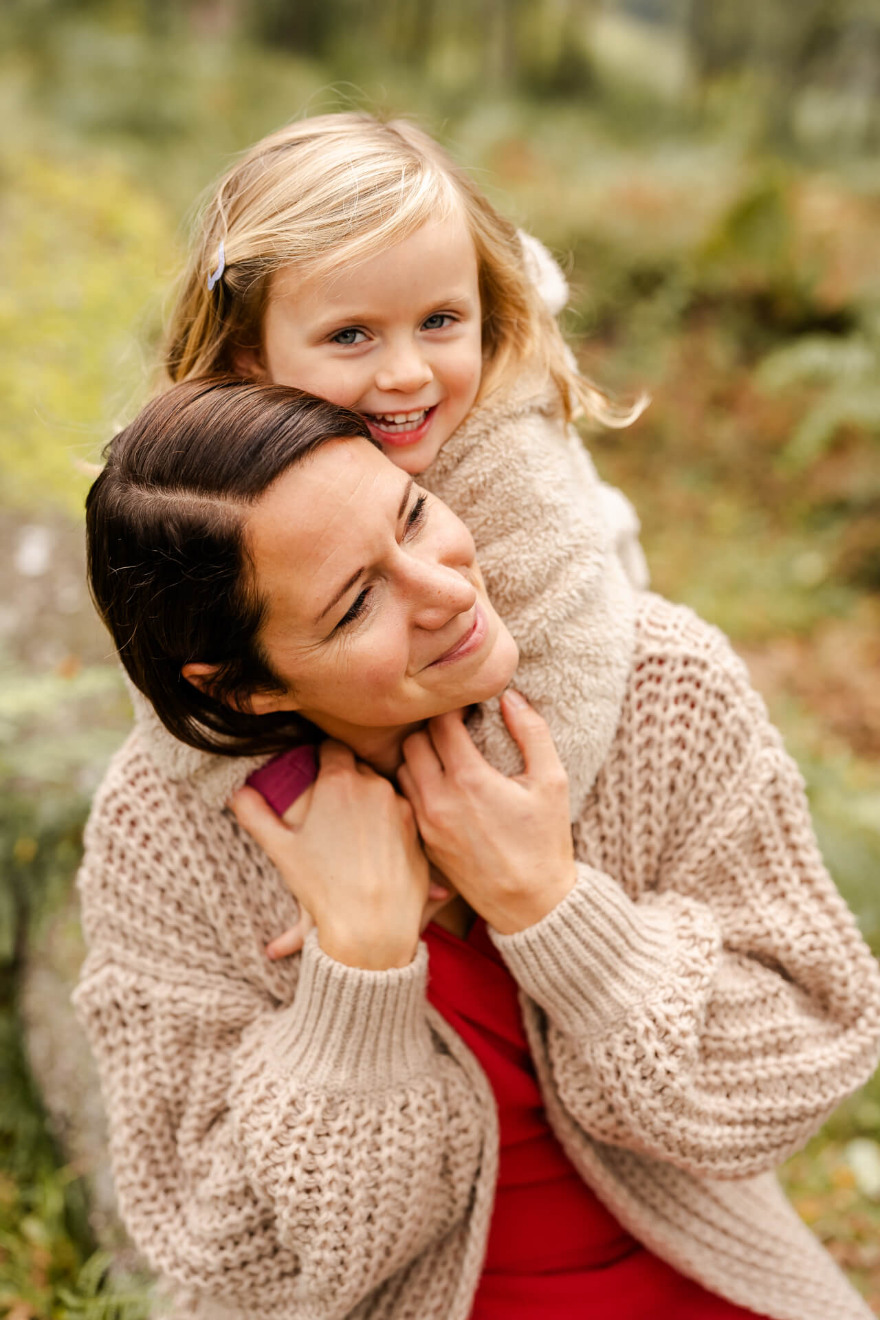 Mama sitzt auf einem Baumstamm, ihre Tochter steht hinter ihr und umarmt sie, beide lachen