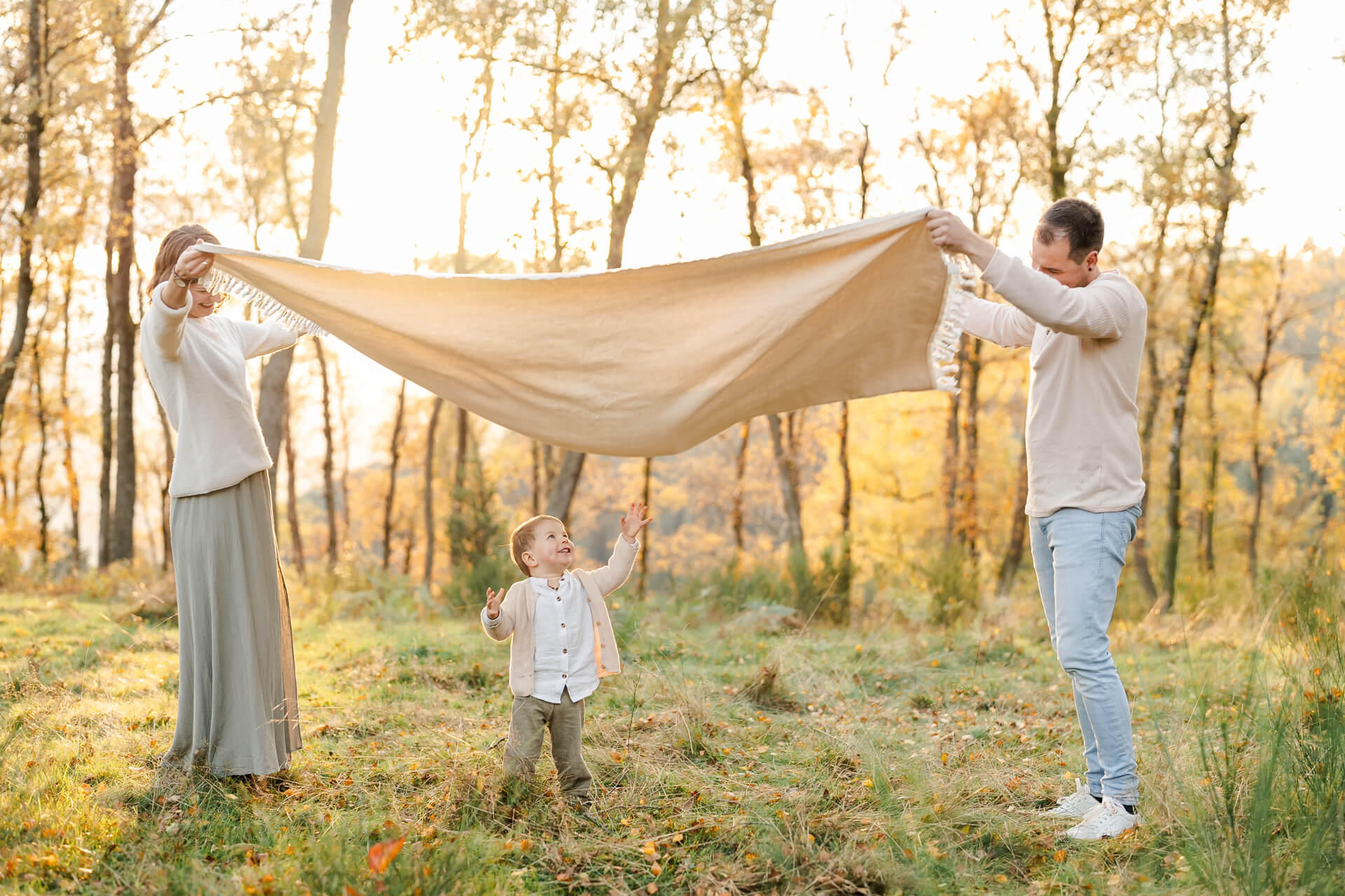 Mama und Papa stehen auf einer Lichtung und schwingen eine Decke in die Höhe, der kleine Sohn steht darunter und lacht