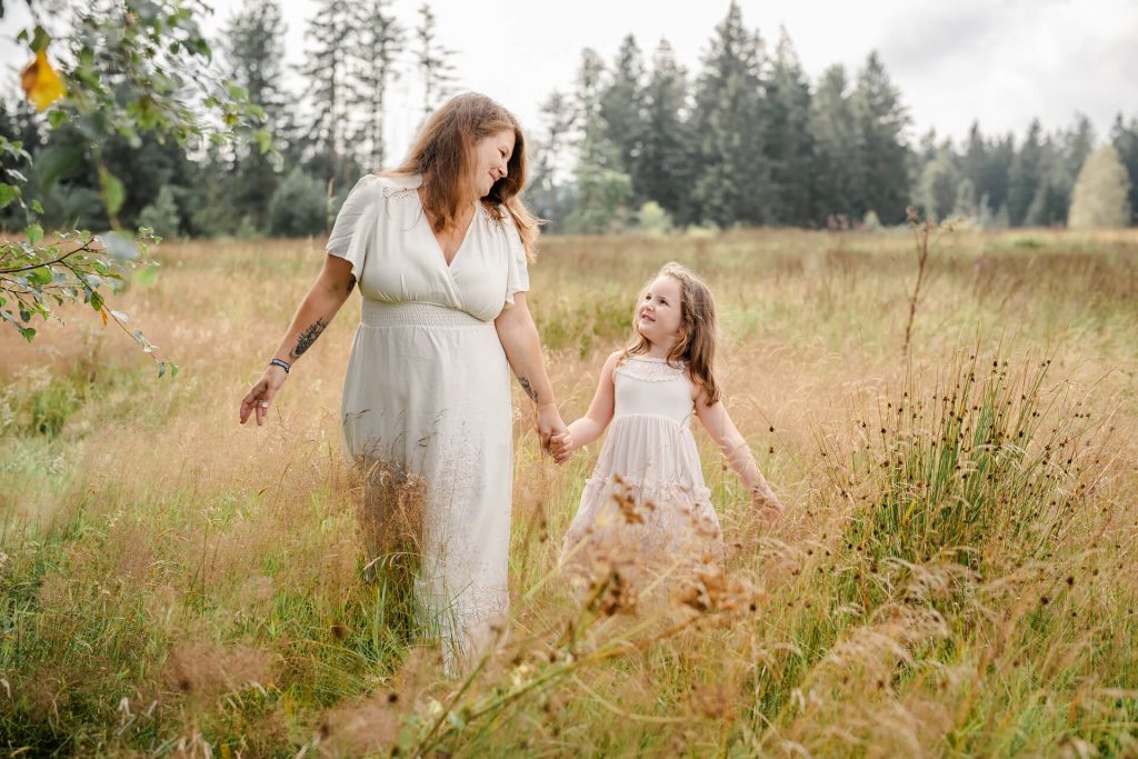 Mama und Tochter laufen Hand in Hand über eine Wiese mit hohem Gras, sie schauen sich an und lachen